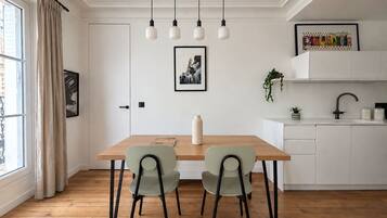 A dining area with a wooden table, two chairs, a hanging light fixture, a framed picture on the wall, and a kitchen area with a sink and cabinets.