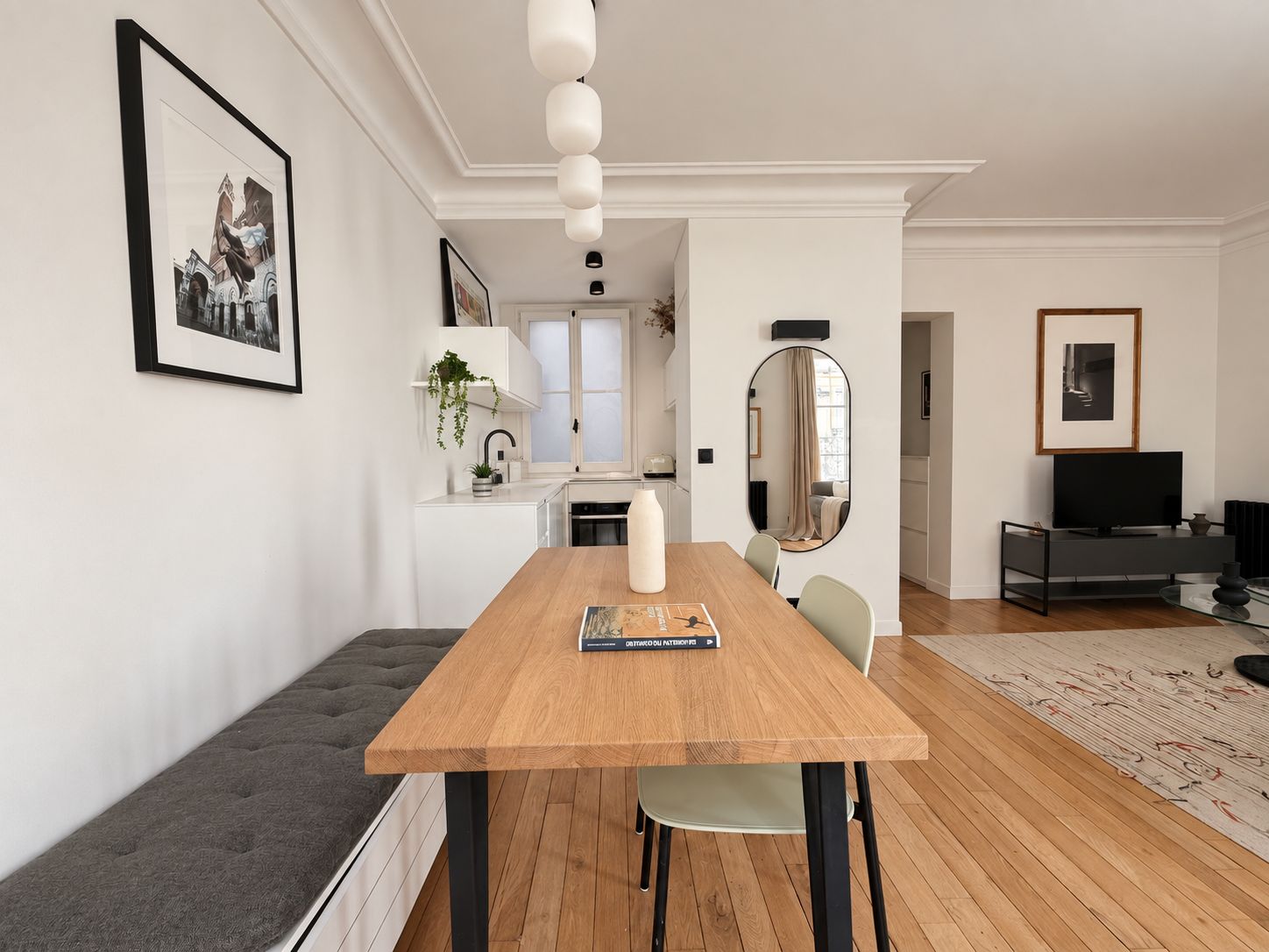 A modern living space with a wooden dining table, a bench, a kitchen area, and a television in the corner.