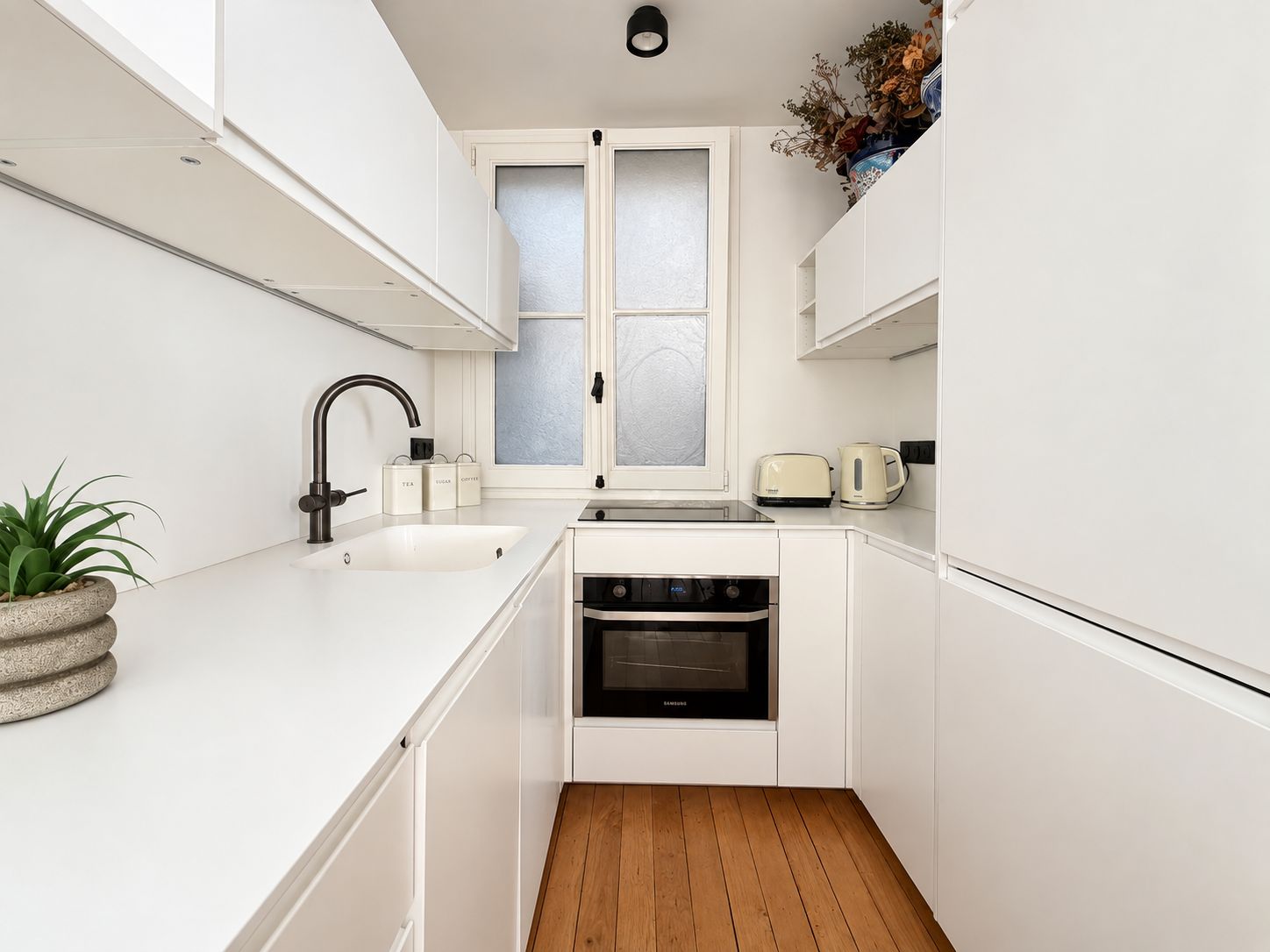 A compact kitchen with white cabinetry, a built-in oven, a sink, and a window with frosted glass.