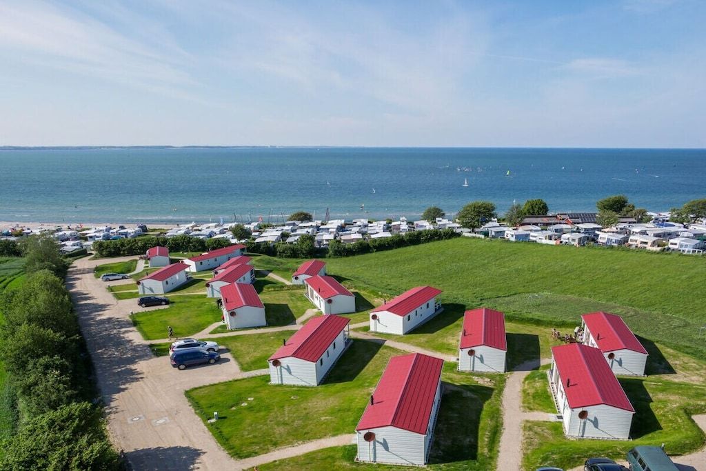 Un camping con cabañas blancas y techos rojos, dispuestas en filas, con un sendero de tierra que conduce a la playa.
