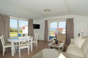 Ein Zimmer mit einem weißen Sofa, einem Esstisch mit Stühlen, einem an der Wand befestigten Fernseher und großen Fenstern mit Vorhängen.