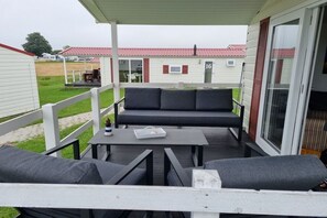 Ein überdachter Außenbereich mit einer Couch und Tischen, einem weißen Zaun und einem Gebäude mit rotem Dach im Hintergrund.