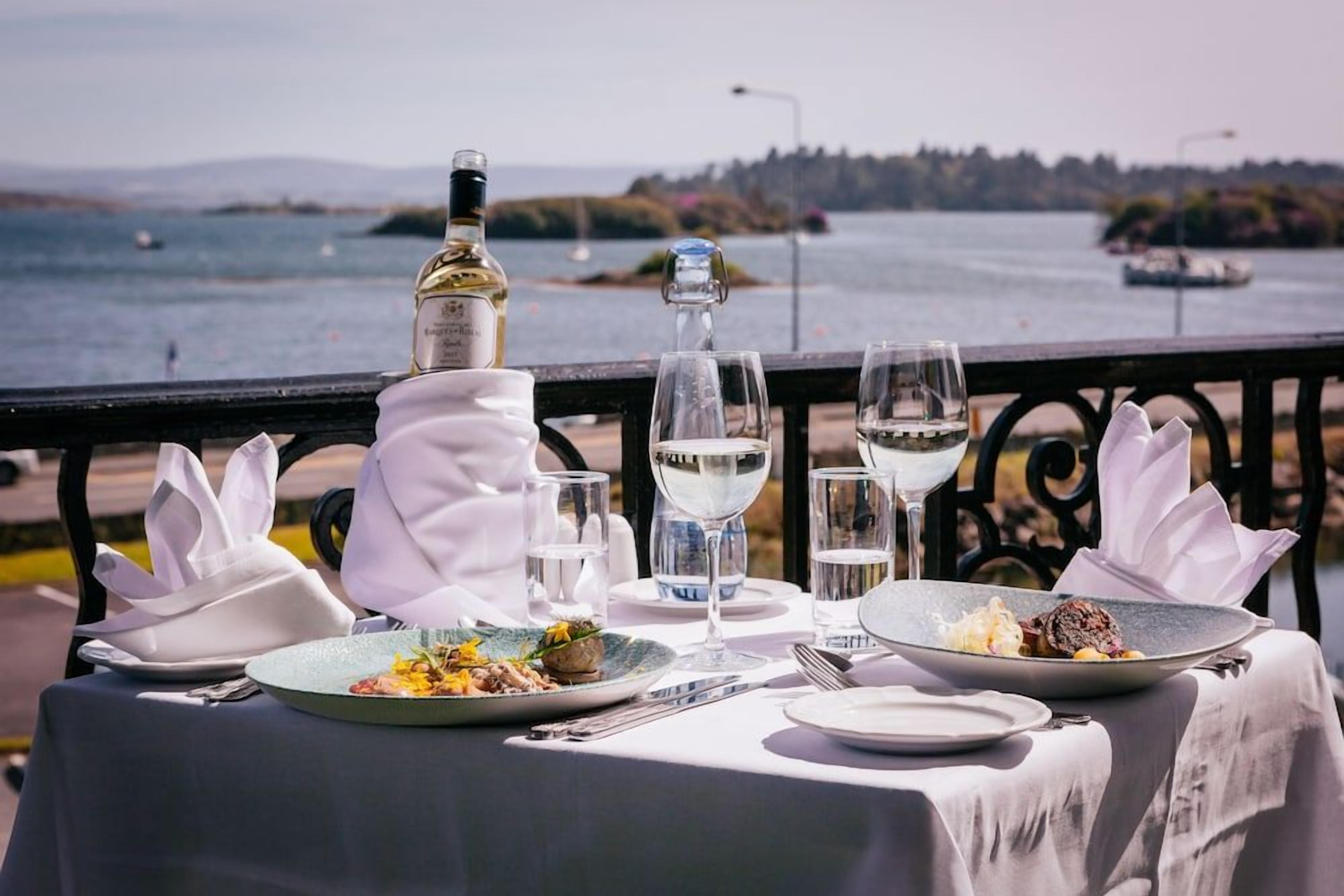 A table set for two with plates of food, glasses, and a bottle of wine, overlooking a body of water.