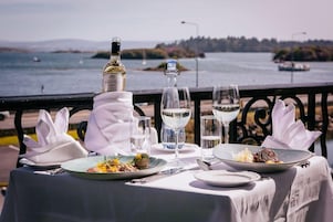 Mesa para dos con platos de comida, vasos y una botella de vino, con vista a un cuerpo de agua.