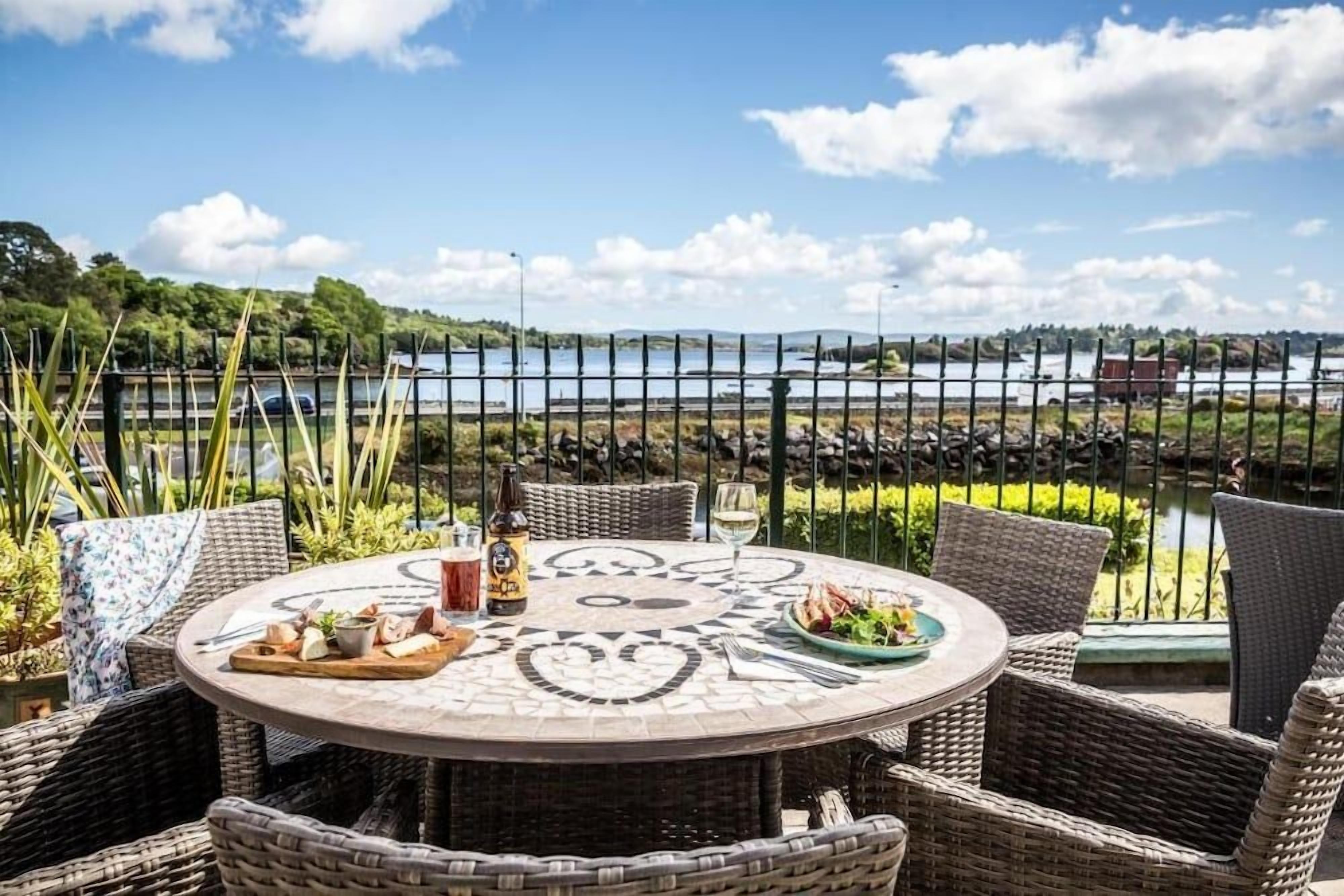 A round table with wicker chairs, a bottle, and a glass of wine, set against a backdrop of a lake and a fence.