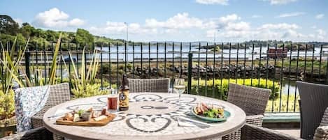 Una mesa redonda con sillas de mimbre, una botella y una copa de vino, frente a un lago y una cerca.