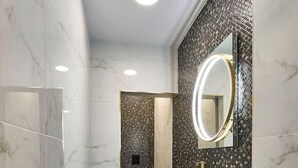 Un baño moderno con tocador de mármol, un espejo redondo y una zona de ducha con azulejos oscuros.