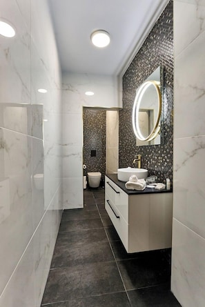 Un baño moderno con tocador de mármol, un espejo redondo y una zona de ducha con azulejos oscuros.