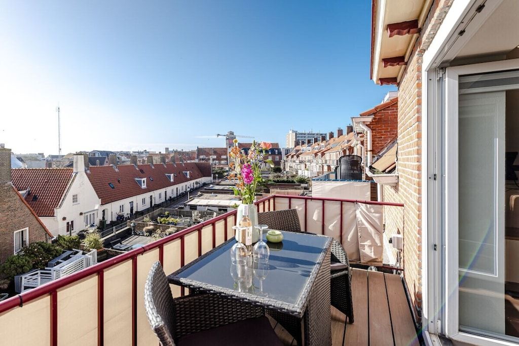 Un balcón con una mesa de vidrio y sillas de mimbre, con vistas a una zona residencial con casas escalonadas.