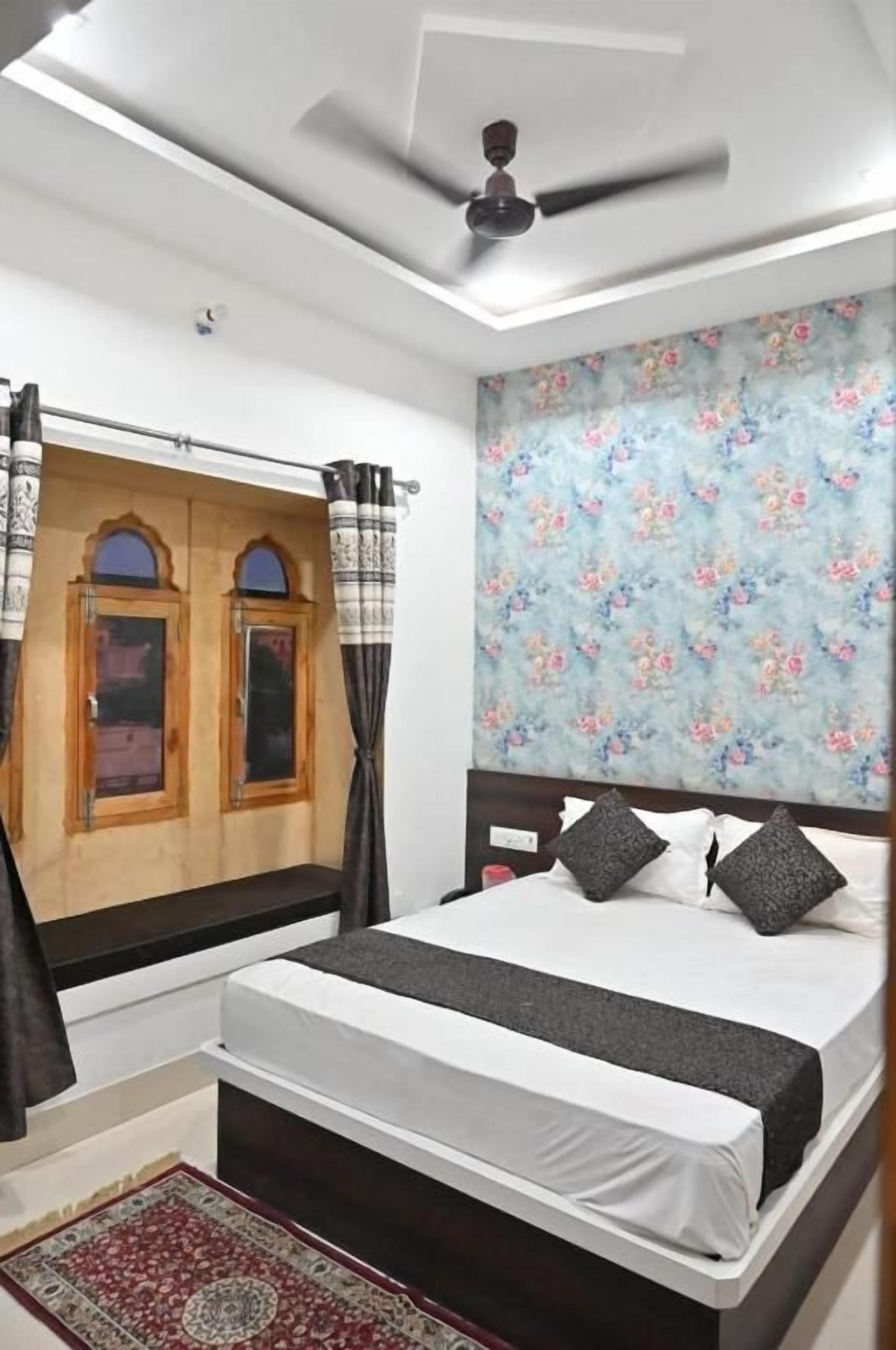 A bedroom with a bed, curtains, a ceiling fan, a floral wallpaper, and a wooden door.