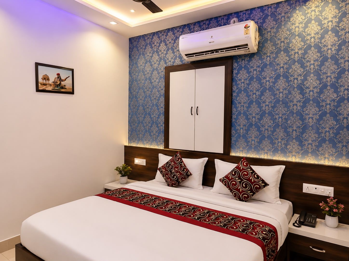 A hotel room with a bed, bedside table, nightstand, and an air conditioning unit.