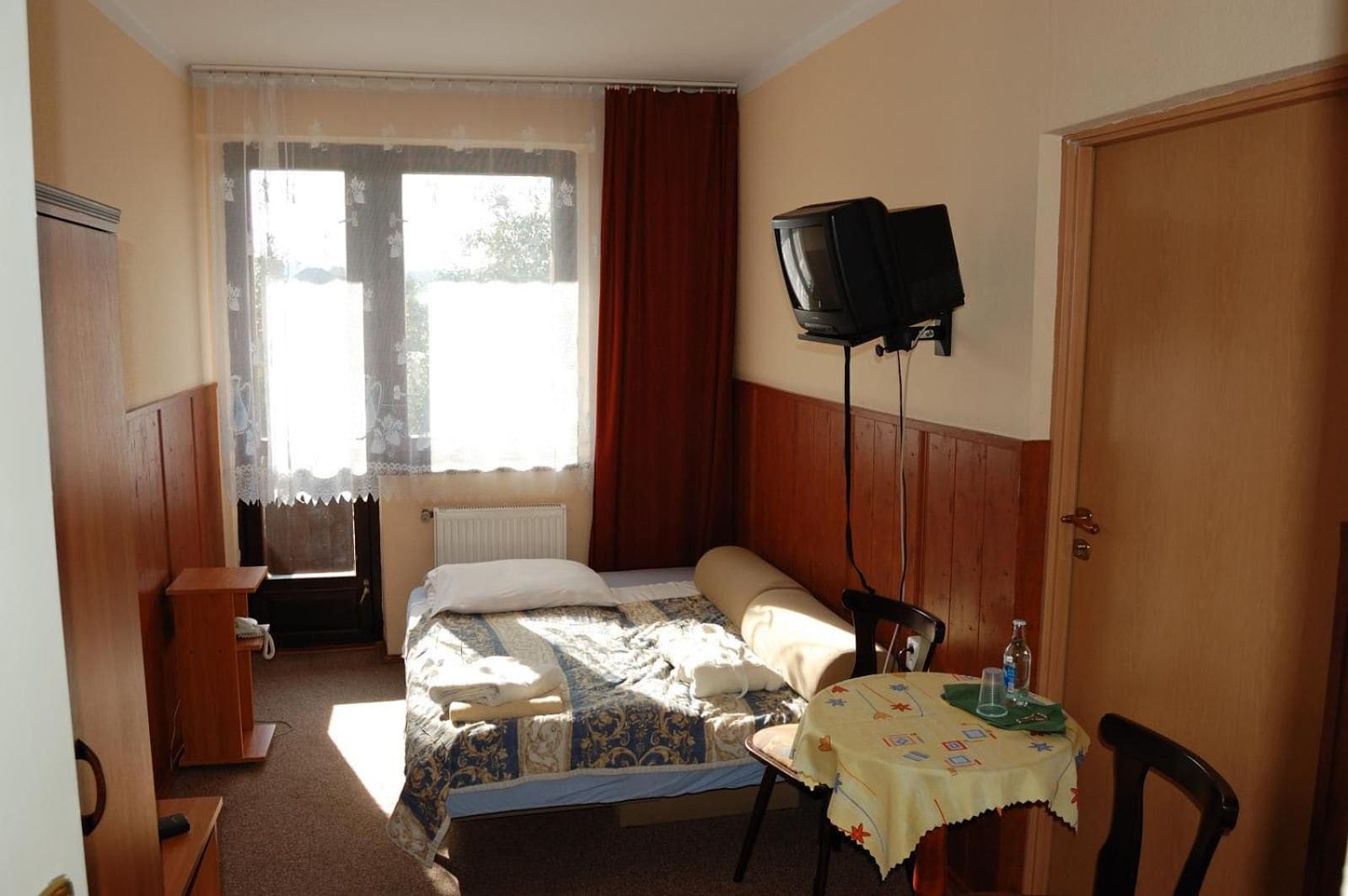 A hotel room with a bed, a television mounted on the wall, a small table with a bottle, and a window with curtains.