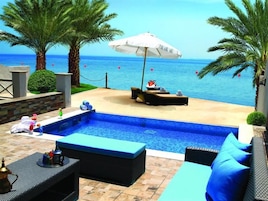 Una piscina con sillones y sombrillas, vista al mar y palmeras.