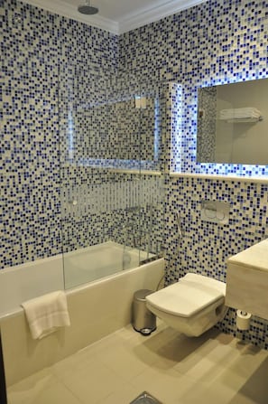 Casa de banho com paredes em mosaico, chuveiro com cabine de vidro, sanita e lavatório.
