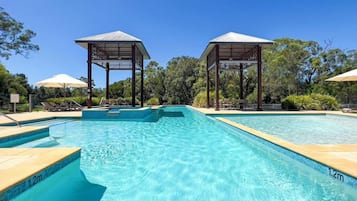 A swimming pool with clear blue water, surrounded by lounge chairs and umbrellas, and two wooden gazebos.
