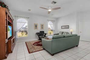 Ein Wohnzimmer mit einem grünen Sofa, einem Fernseher, einem Deckenventilator und einem Fenster mit Blick ins Freie.