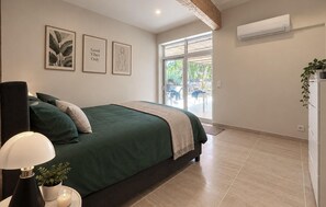 A bedroom with a bed, a bedside table with a lamp, a potted plant, and a window with a view of the outdoors. - Maison piscine au calme (Méjannes-Le-Clap)