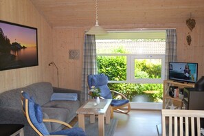 Ein gemütliches Wohnzimmer mit einer Couch, Sesseln, einem Fernseher und einem Fenster mit Blick auf Grünflächen.