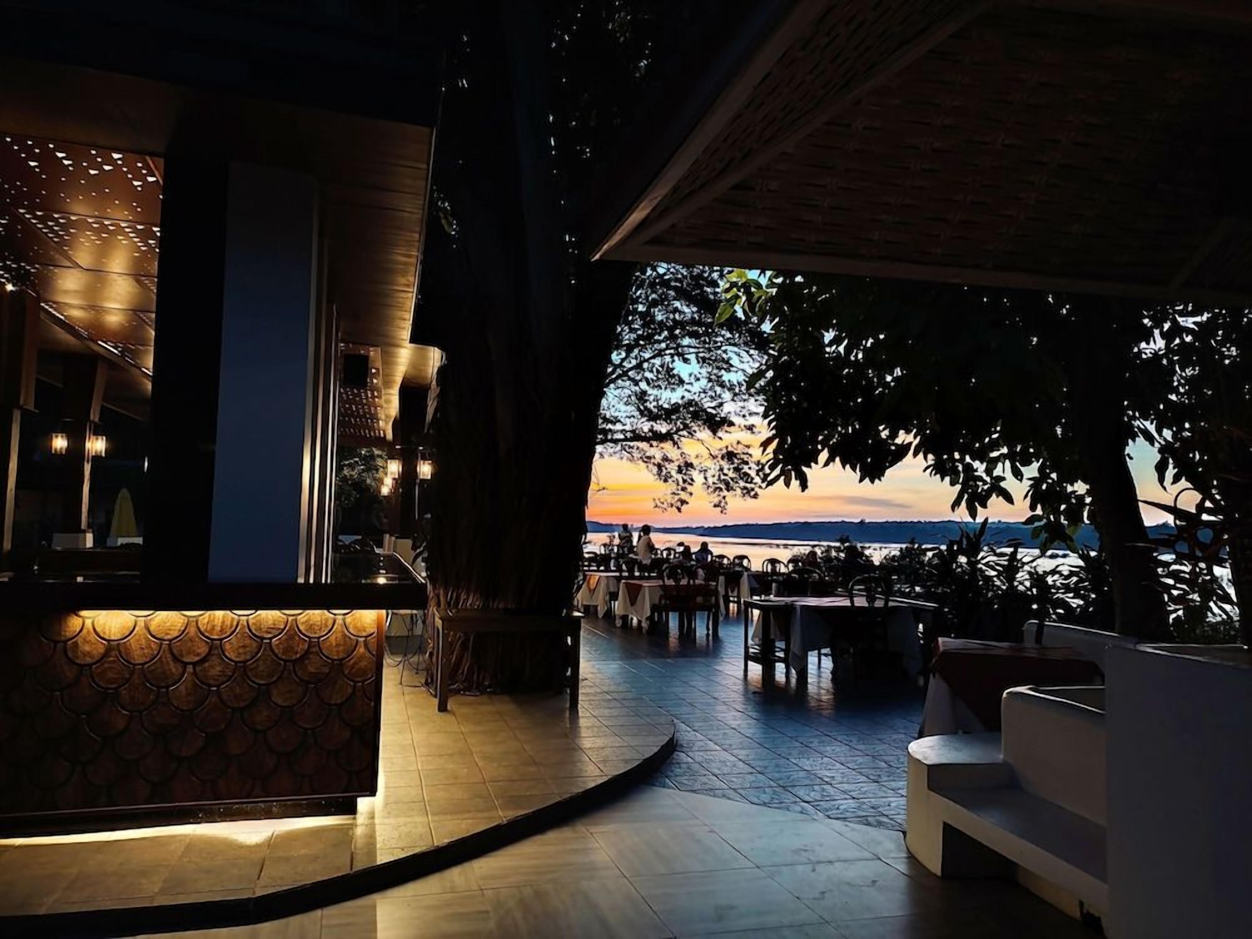 An outdoor dining area with tables and chairs, a reception desk, and a view of the sunset over a body of water.