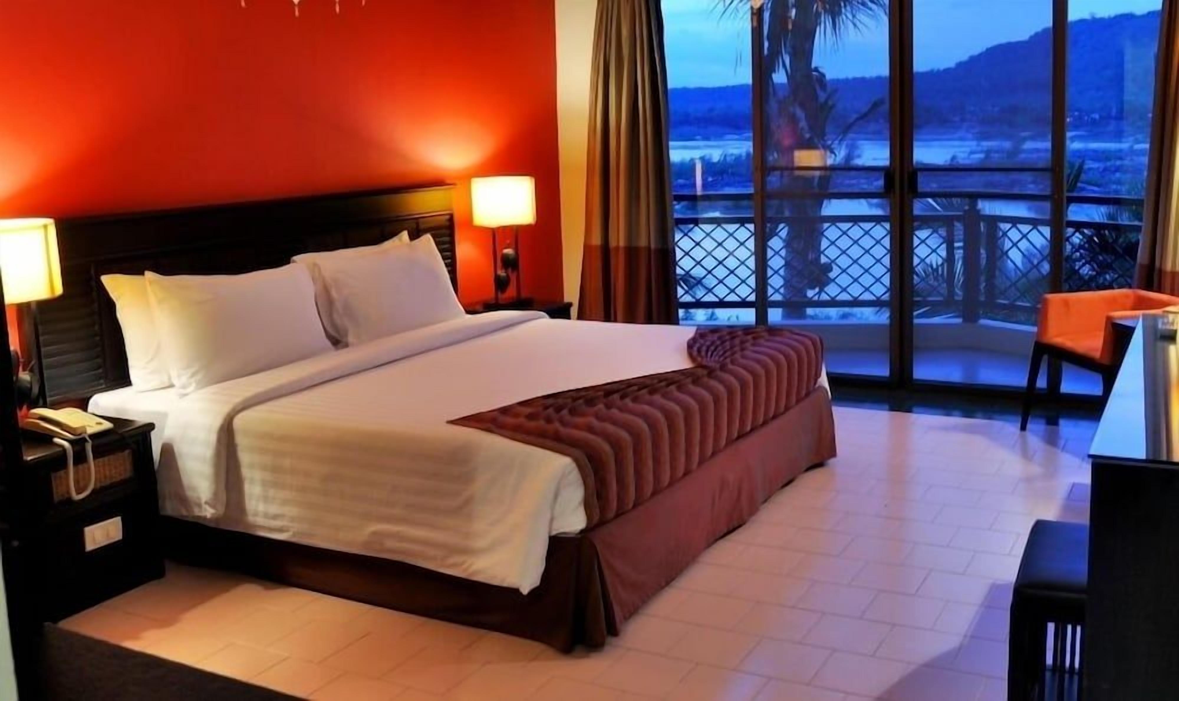 A hotel room with a bed, a nightstand, a lamp, a view of the outdoors, and a balcony with a chair.