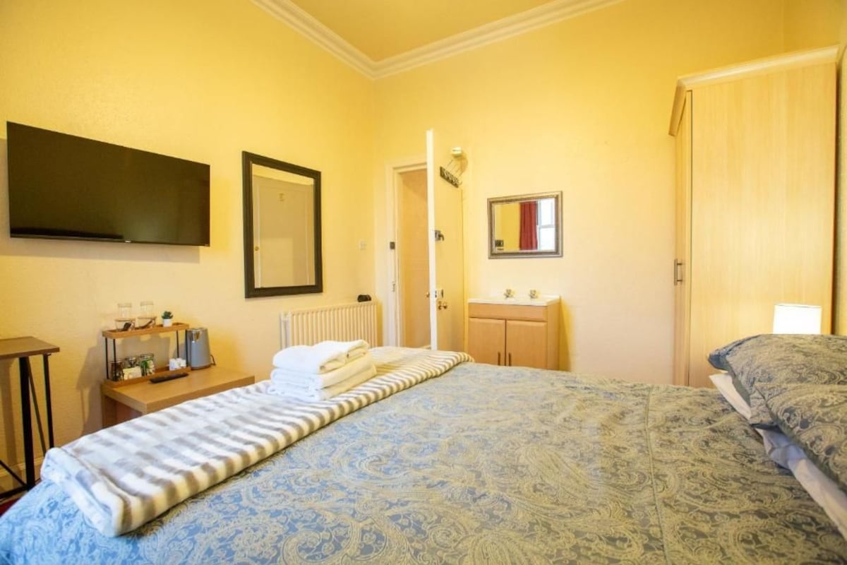 Une chambre d’hôtel avec un lit, une table de chevet avec une lampe, un téléviseur fixé au mur et une porte donnant sur une autre pièce.