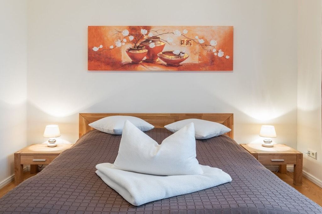 Un lit bien fait, recouvert d’une couverture marron à texture et agrémenté d’oreillers blancs, avec deux tables de chevet dotées de lampes de chaque côté, et un tableau de fleurs accroché au mur.