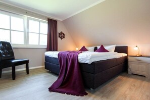 A bedroom with a bed, a chair, a nightstand, a lamp, a window with curtains, and a wall decoration. - Holiday home with 2 bedrooms (Fuhlendorf)
