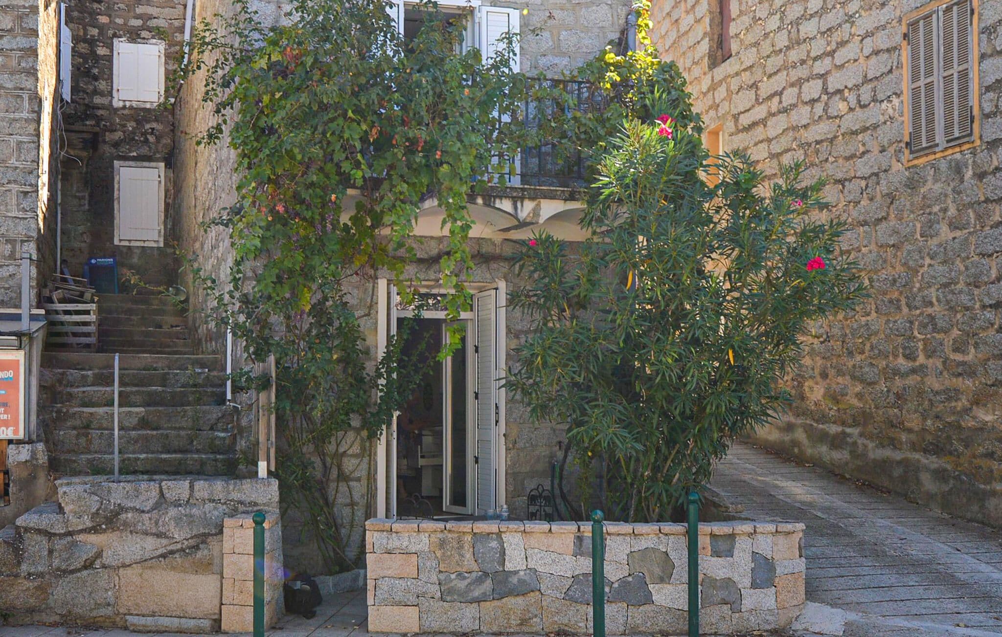 A stone building with a staircase leading to a door, surrounded by greenery and a stone wall.