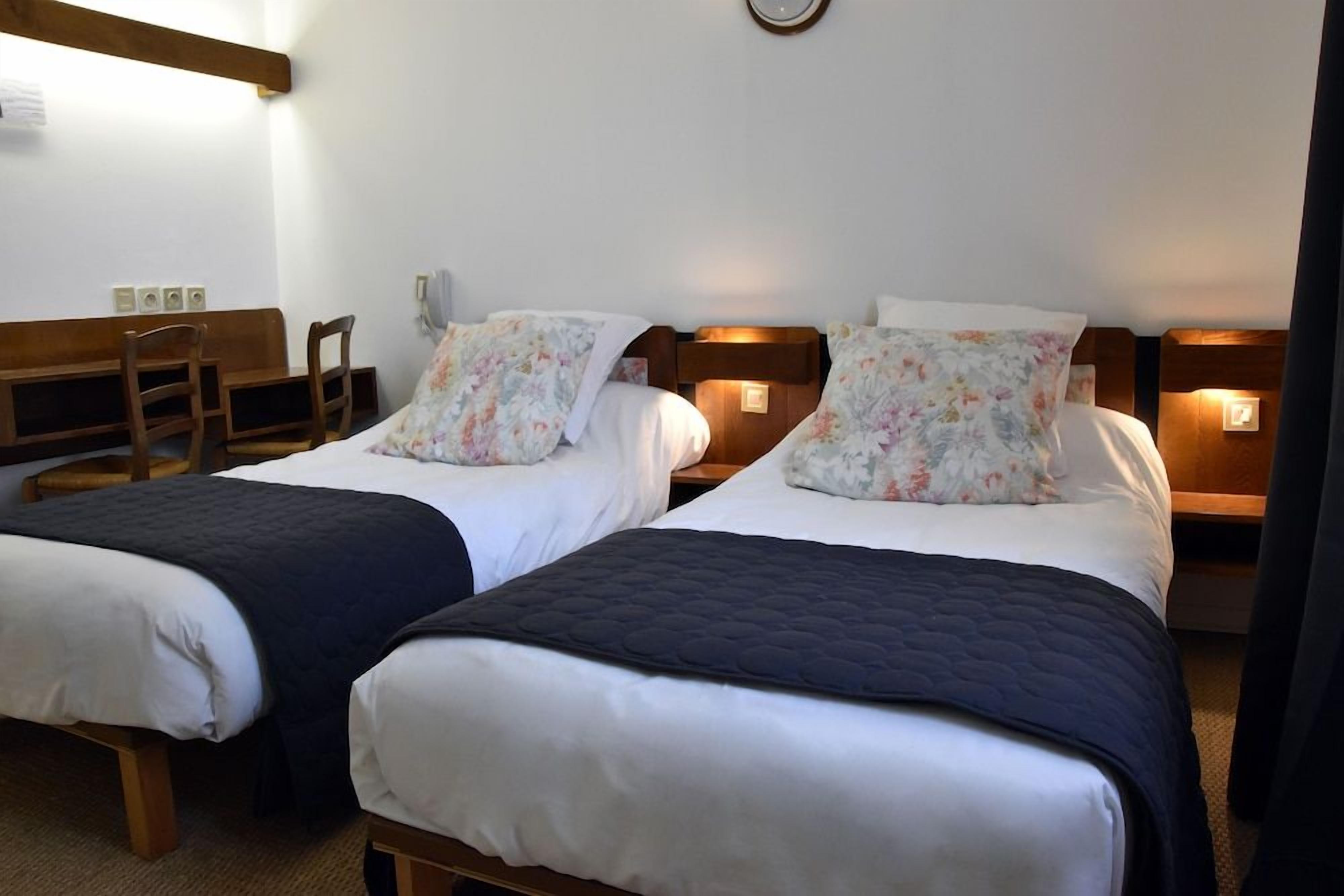 Une chambre d’hôtel avec deux lits simples, chacun doté d’une tête de lit en bois, de draps blancs et d’un coussin à motifs floraux. On y trouve des tables de chevet avec des lampes, un banc en bois et une horloge murale.