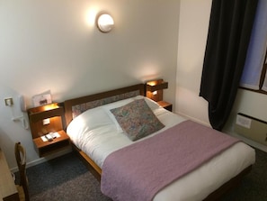 Cama bem arrumada com edredom branco e manta roxa, acompanhada por duas mesas de cabeceira com abajures.