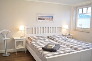 Een slaapkamer met een wit bed, geruit beddengoed, bedlampen, een nachtkastje, een lijst met een foto aan de muur en een raam met jaloezieën.