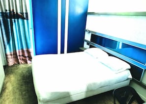 A single bed with a white mattress and pillows, a blue wall, and a window with curtains. - Enzo Trappes Versailles (Trappes)