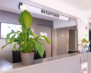 A reception area with a reception desk, a potted plant, and a sign above the desk that reads 'RECEPTION'. - Enzo Trappes Versailles (Trappes)