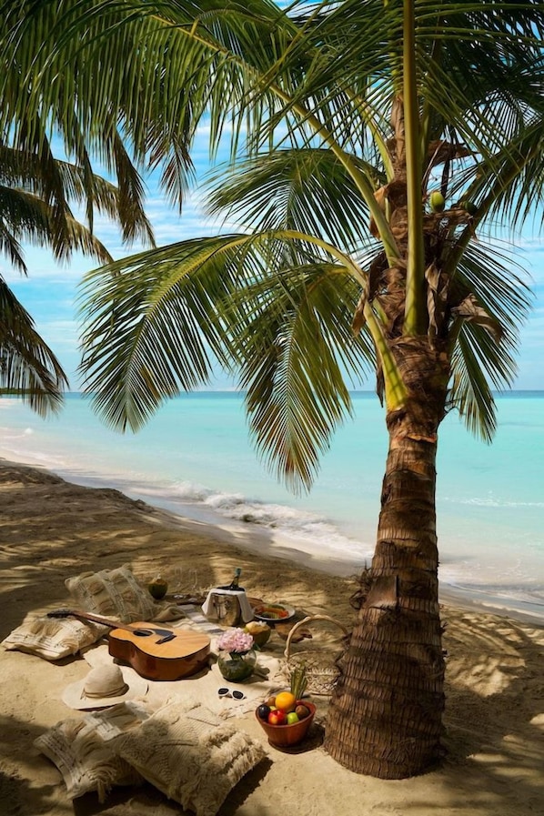 A beach scene with a palm tree, a guitar, a hat, a bowl of fruit, and a blanket on the sand. - Bangaram Island - Ihcl Seleqtions (Bangaram Island)