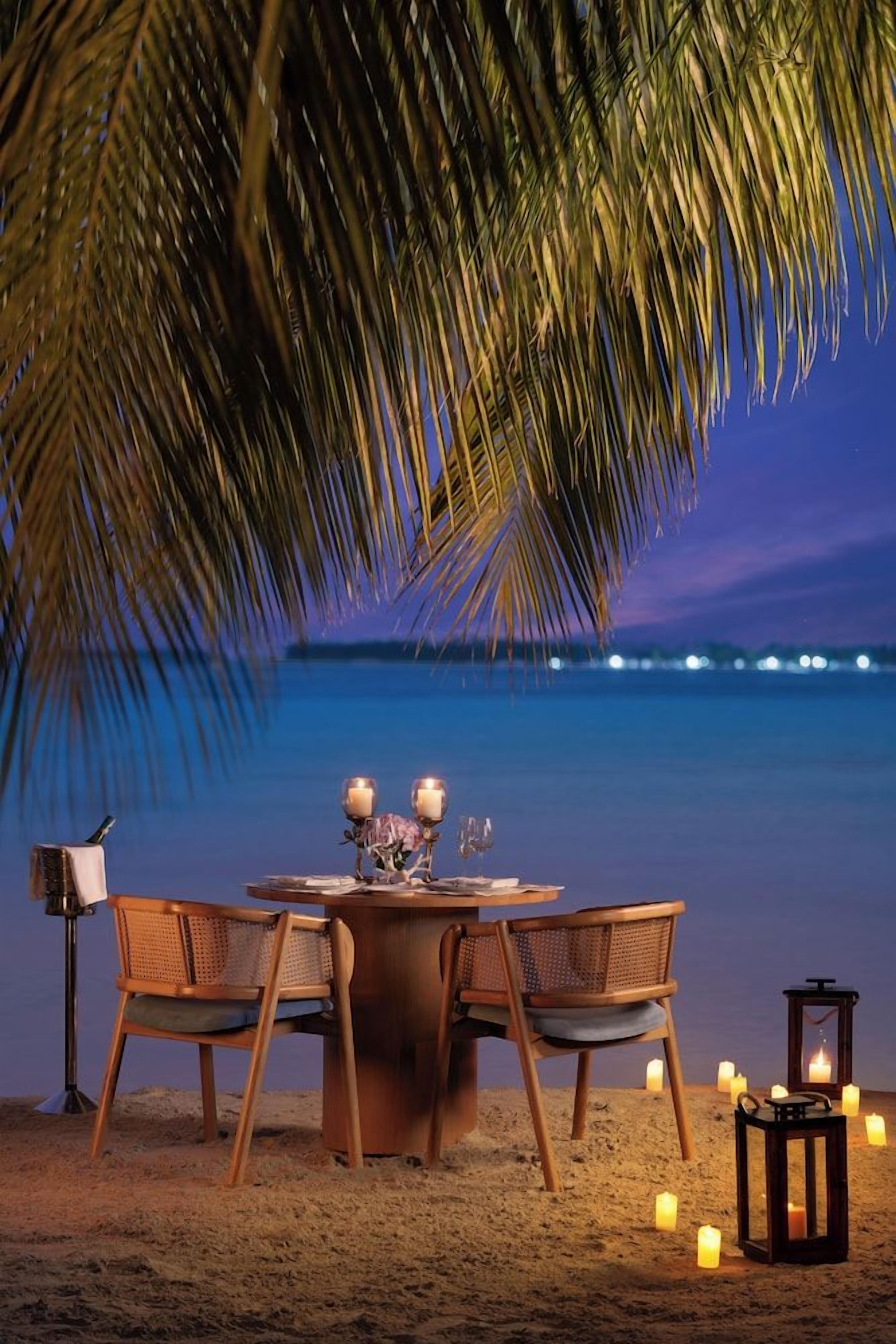 A beachside dining setup with a table, chairs, candles, and a lantern.