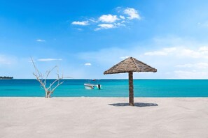 Una playa con una sombrilla de paja, agua turquesa cristalina y un barco a lo lejos.