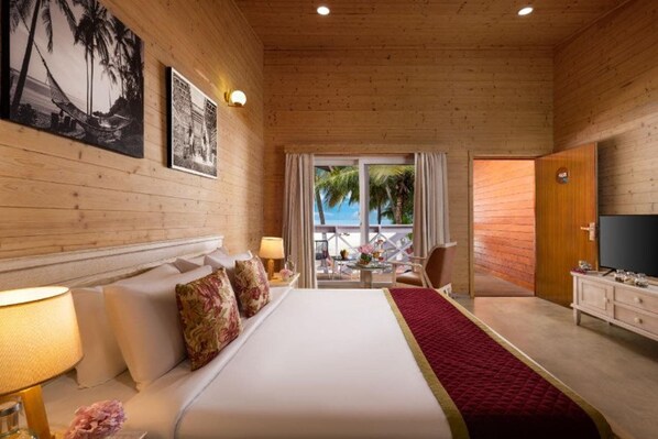 A hotel room with a bed, a TV, a desk with a chair, and a window with a view of palm trees. - Bangaram Island - Ihcl Seleqtions (Bangaram Island)
