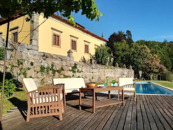 Una terraza de madera con mobiliario exterior, piscina y un edificio de paredes de piedra.