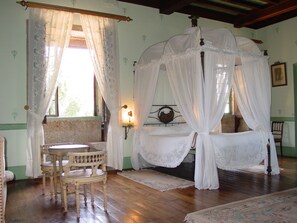 Quarto com cama de dossel, uma cadeira de madeira, uma mesinha e um criado-mudo.