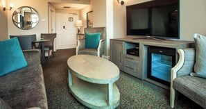 Ein Hotelzimmer mit Sitzgelegenheit, Fernseher, Minibar und einer Tür zu einem Flur.