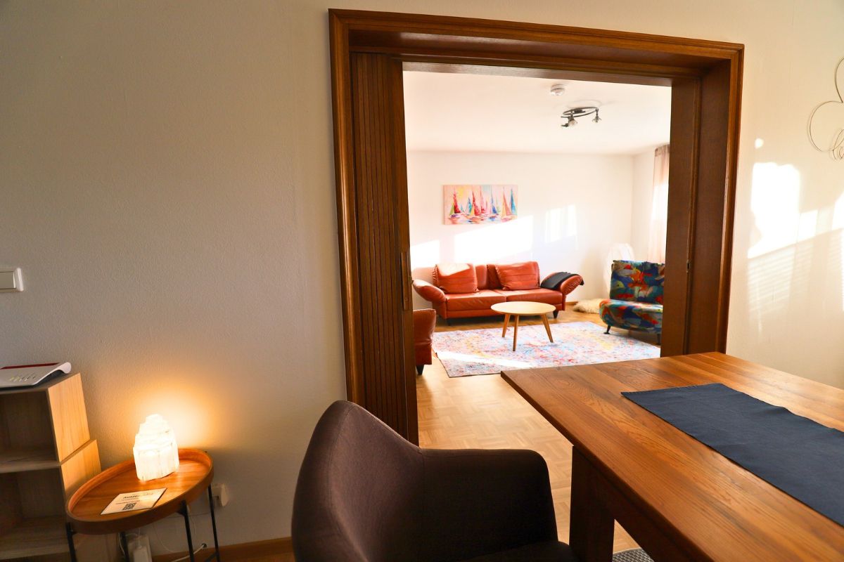 Ein Zimmer mit einem Holztisch, einem Stuhl, einem kleinen Tisch mit Lampe und einer Couch im Hintergrund.