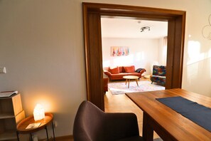 Ein Zimmer mit einem Holztisch, einem Stuhl, einem kleinen Tisch mit Lampe und einer Couch im Hintergrund.