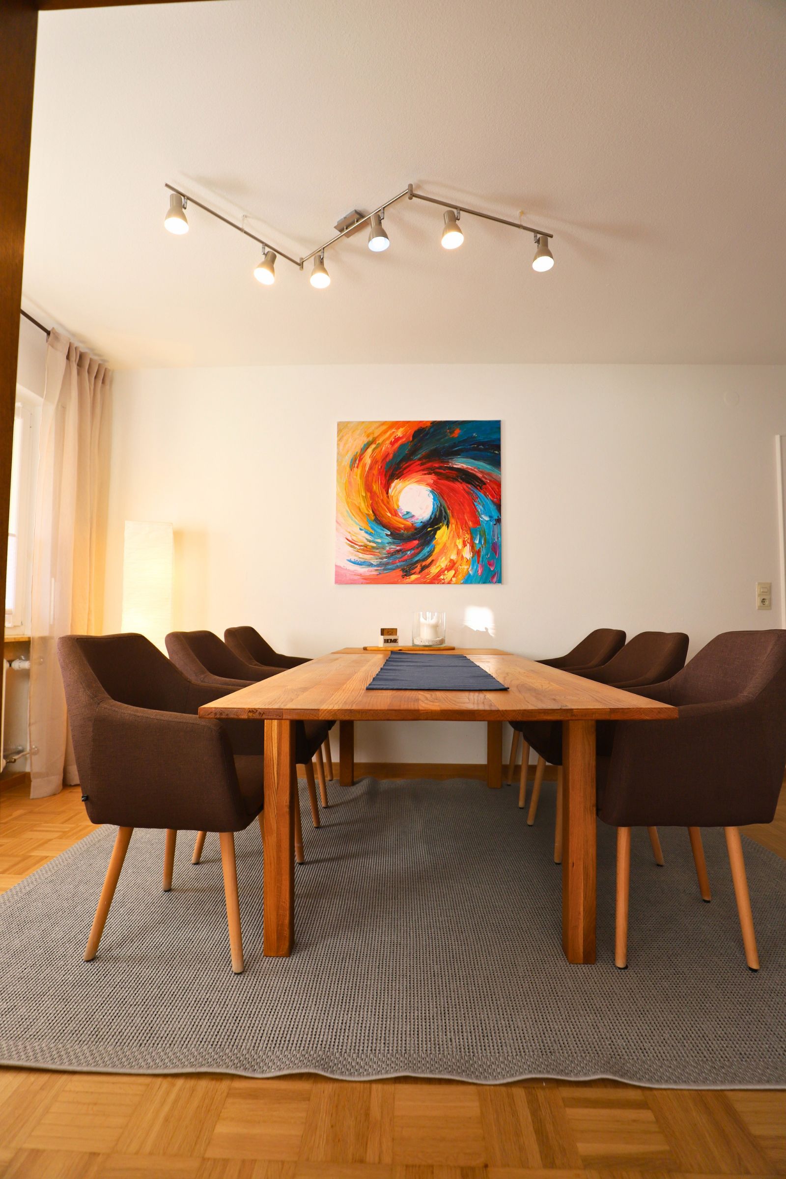 Ein Konferenzraum mit einem Holztisch, acht Stühlen, einem farbenfrohen abstrakten Gemälde und einer Deckenleuchte.