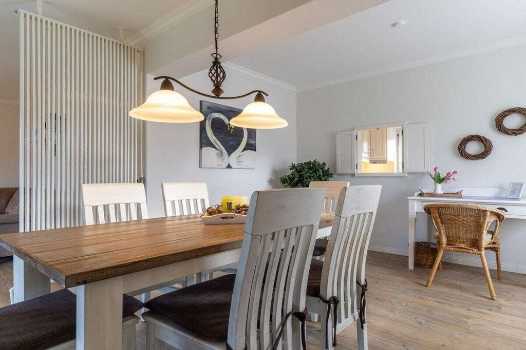 Et spiseområde med et træbord og stole, en hængende lampe samt en hvid væg med et indrammet kunstværk.