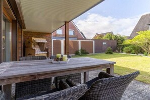 Ein überdachter Außenbereich mit Tisch und Korbstühlen, einer Ziegelwand und Blick auf ein Wohngebäude im Hintergrund.