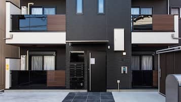 A modern building with a black facade, two balconies, a door with a keypad, and a small outdoor area with a trash bin.