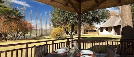 An outdoor dining area with a table set for four, surrounded by a wooden fence, and a thatched roof structure in the background.