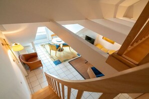 Una habitación con techos altos, una escalera de madera, una zona de comedor con mesa y sillas, y un televisor.