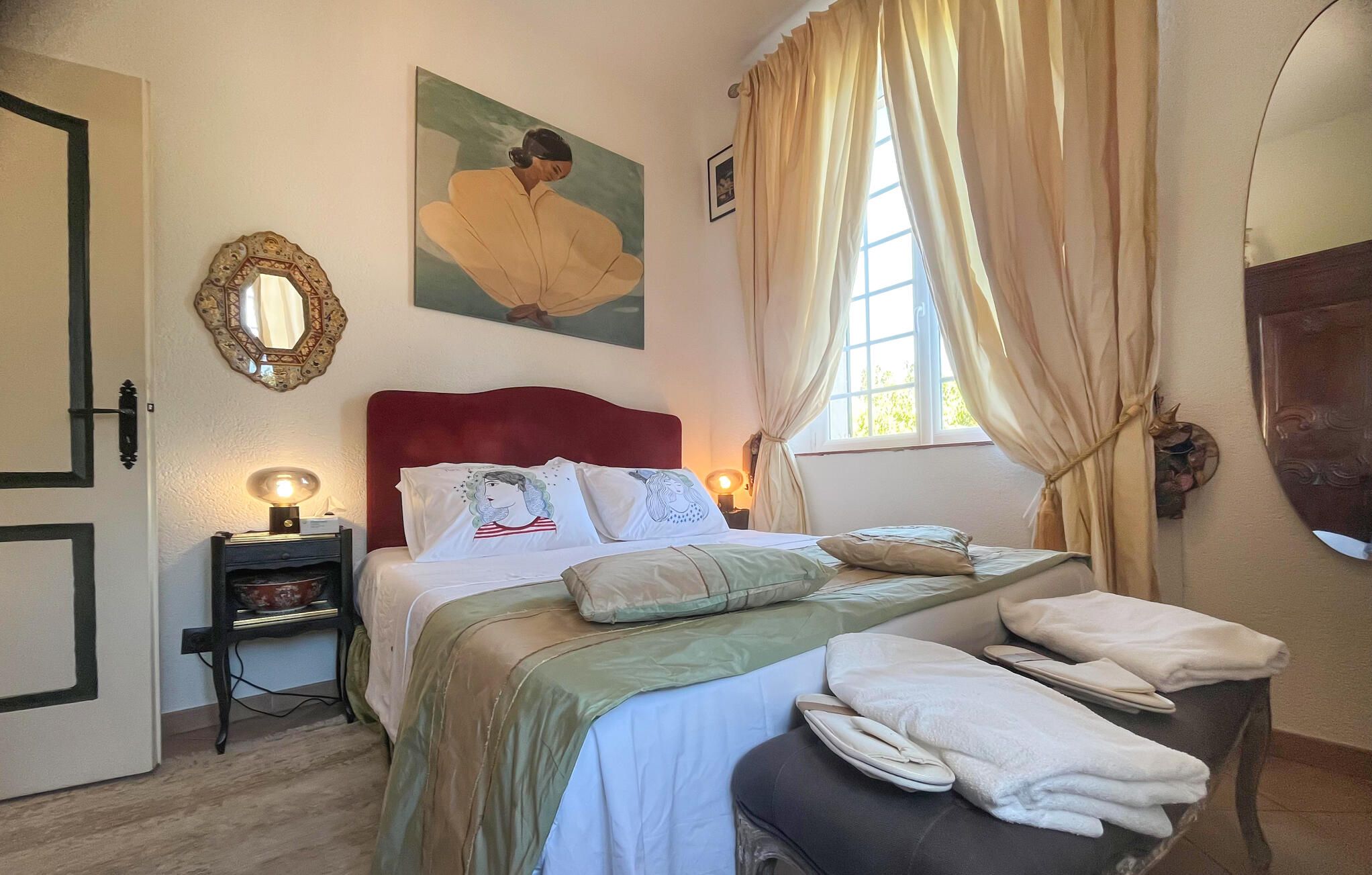 A bedroom with a bed, a bedside table, a lamp, a window with curtains, and a wall-mounted mirror.