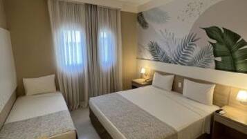 Quarto de hotel com duas camas, uma janela grande com cortinas, um mural na parede e uma mesa de cabeceira com abajur.